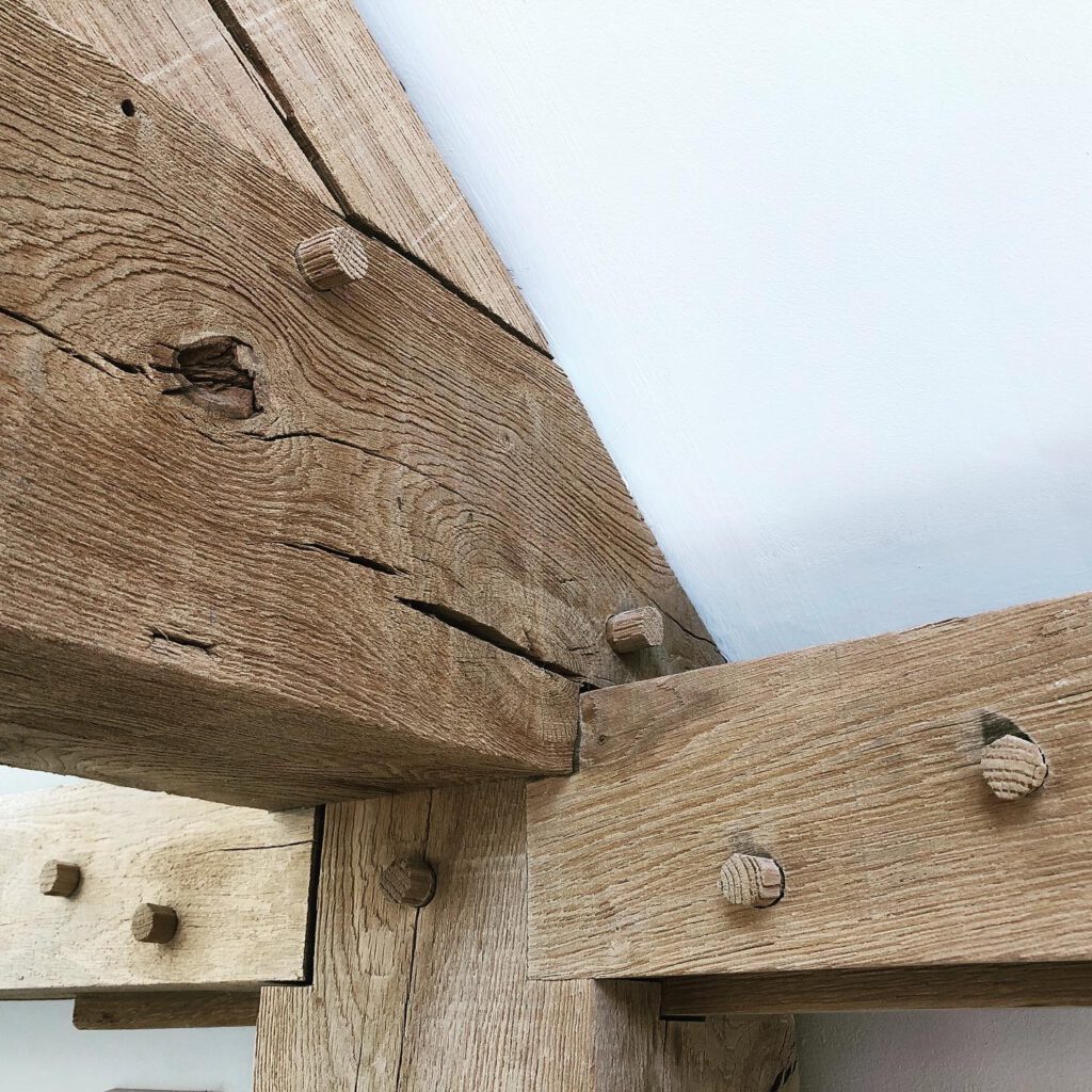 Close-up of rustic wooden beams joined together with dowels, showing the wood’s natural grain, texture, and cracks against a white wall ceiling.