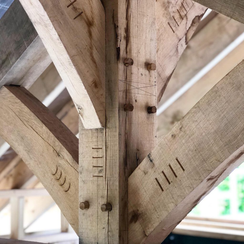 Close-up of wooden beams joined with traditional mortise and tenon joinery, featuring wooden pegs and visible chisel marks, in the framing of a building under construction. Natural light highlights the wood grain.