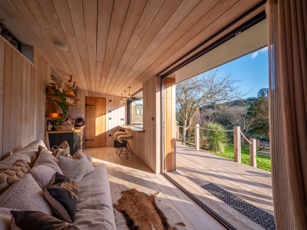 A cozy wooden interior with a sofa, pillows, and a kitchen area opens up to a sunny patio and lush green garden through large glass doors, offering a scenic view of trees and hills.