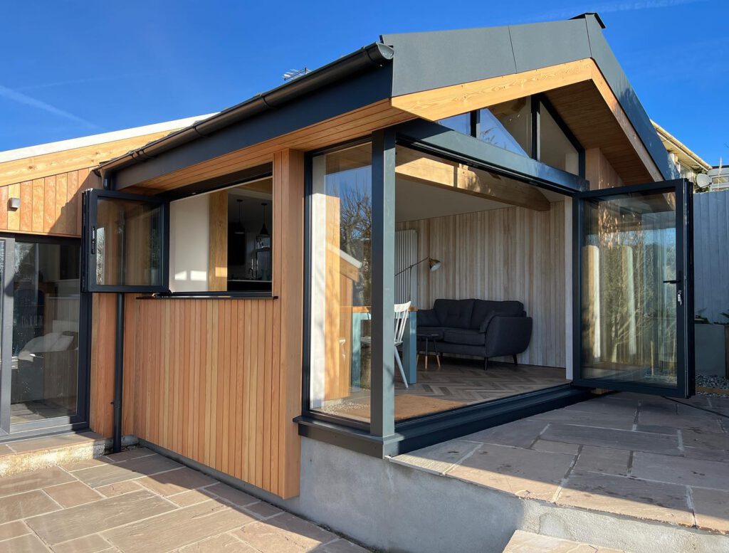 A modern house with wood and glass exterior, large folding glass doors open to a living area with a gray sofa and wooden walls, under a clear blue sky.
