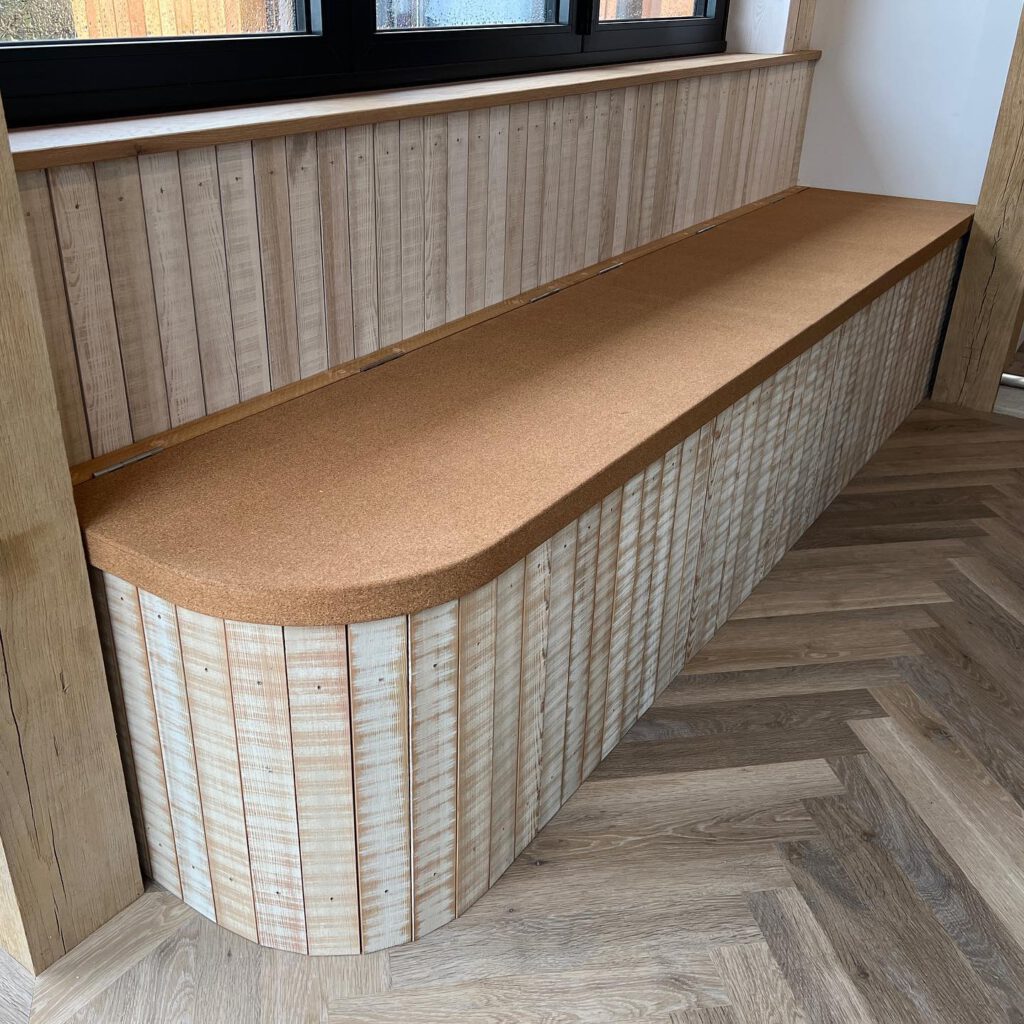 A wooden bench with a cork seat and vertical slat paneling, set against a wall with a window above. The bench sits on a herringbone-patterned wooden floor.