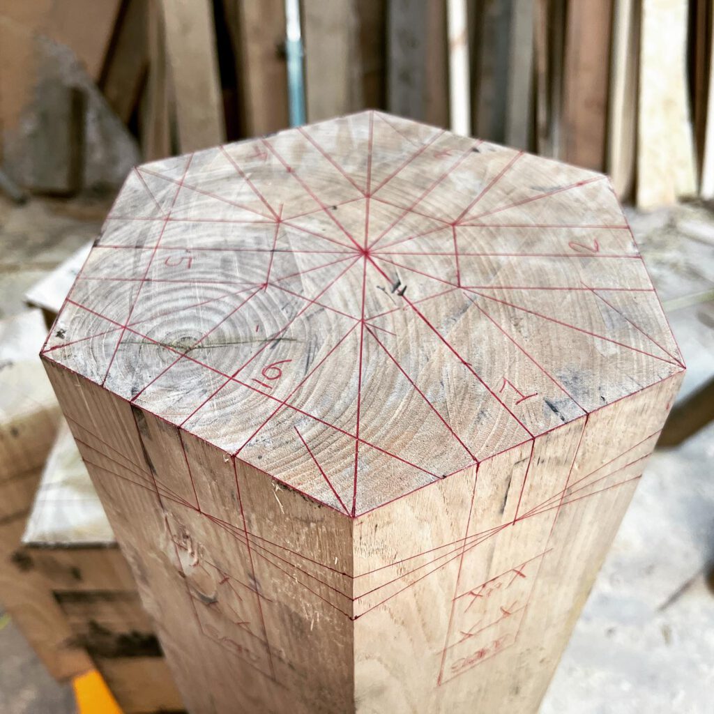 A hexagonal wooden post marked with red guide lines and numbers, likely for precise cutting or carving, stands upright in a workshop with various wooden boards in the background.