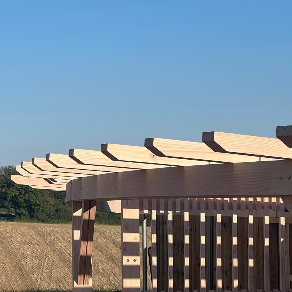 A close-up side view of a wooden pergola under construction, with beams and rafters visible, set against a clear blue sky and open grassy field in the background.