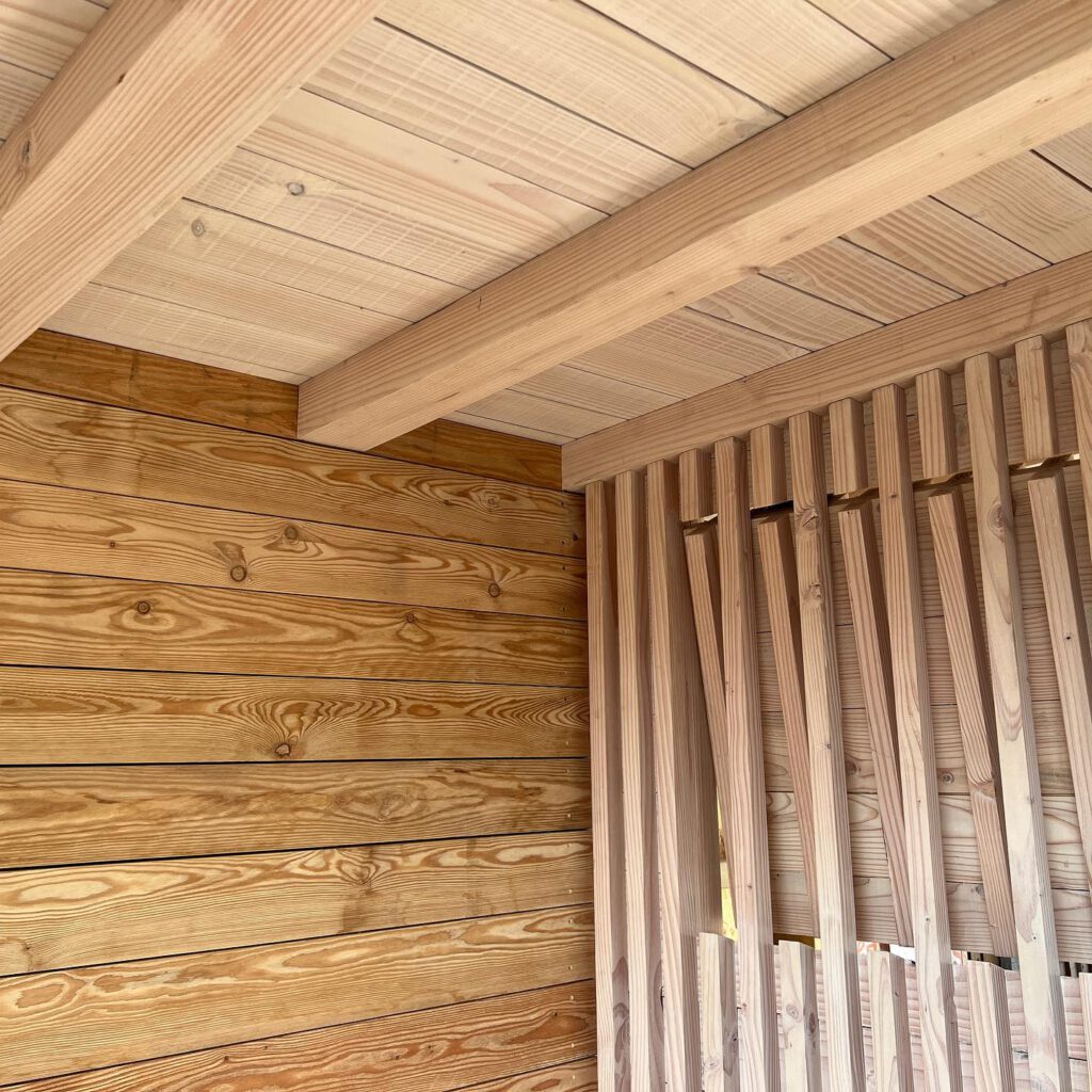 Interior view of a wooden structure showing a ceiling with beams, horizontal wooden wall panels, and a partially slatted vertical wall on the right side. The wood appears smooth and unfinished.