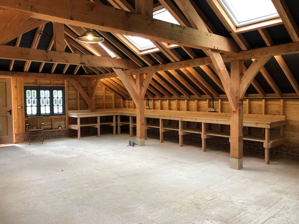 A spacious wooden workshop with exposed beams, skylights, long wooden workbenches along the walls, concrete floor, and a door with small windows. The area is well-lit and organized.
