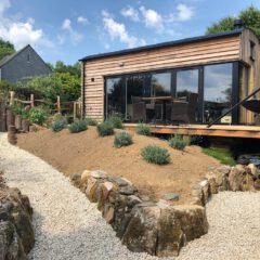 A small modern cabin with wood siding and large glass doors sits next to a deck with outdoor furniture, overlooking a landscaped garden with gravel paths, rock borders, and shrubs under a blue sky.