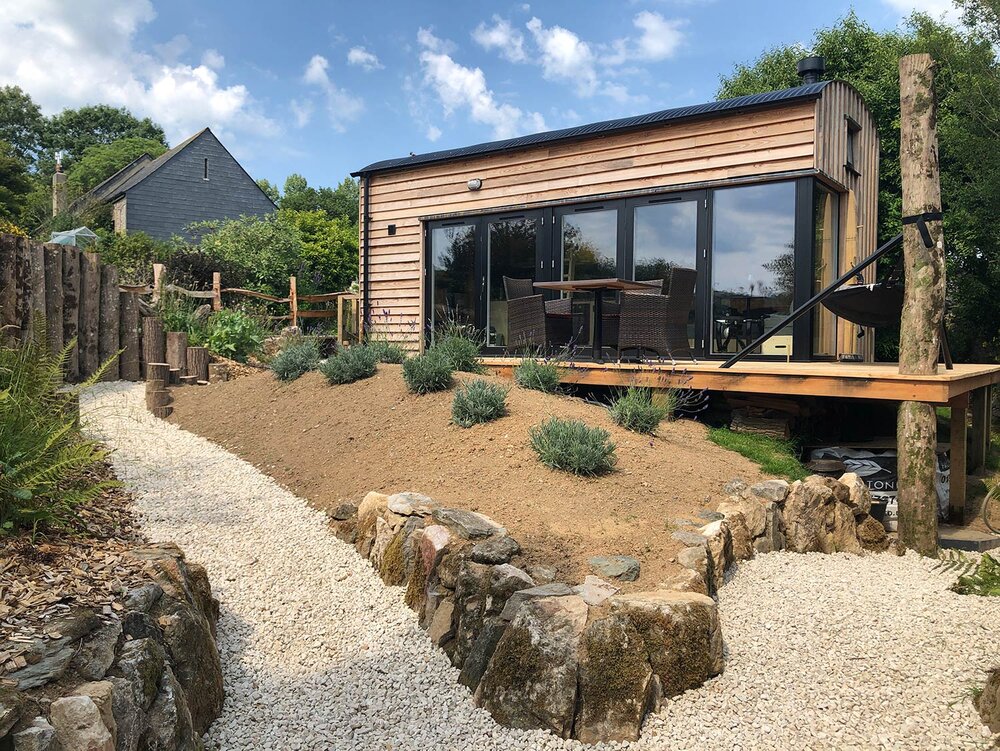 A small modern cabin with wood siding and large glass doors sits next to a deck with outdoor furniture, overlooking a landscaped garden with gravel paths, rock borders, and shrubs under a blue sky.