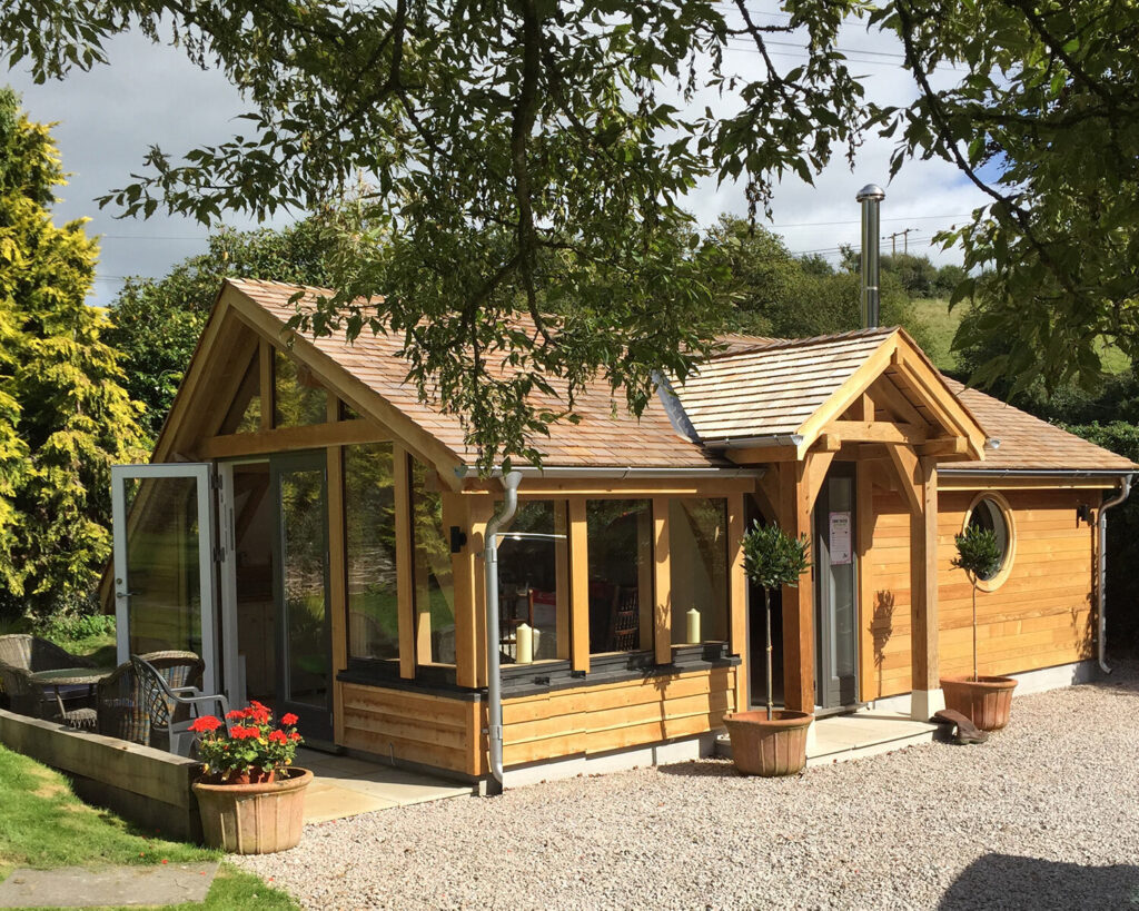 A small modern wooden cabin with large glass windows and doors, surrounded by potted plants and trees, sits on a gravel path with a lush, green landscape in the background.