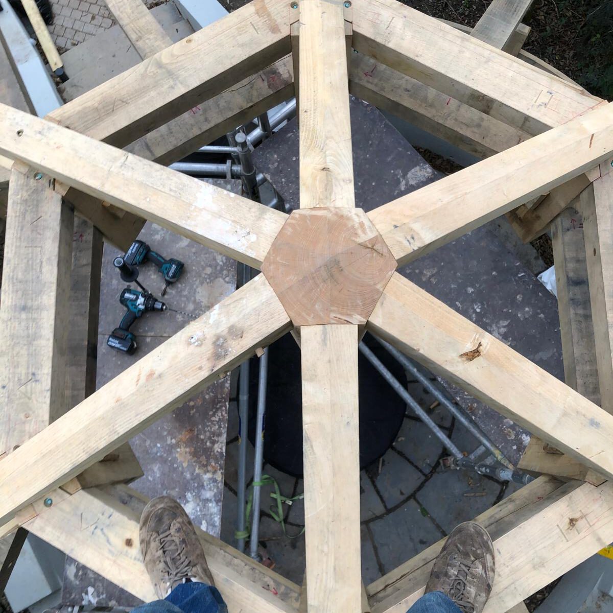 A person standing on a wooden hexagonal structure under construction, viewed from above. Power tools lie on the ground below, and scaffolding is visible around the area.