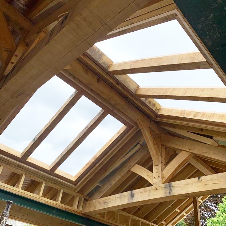 A view from below of a wooden roof structure under construction, featuring two rectangular skylight openings with exposed beams and a cloudy sky visible through the gaps.