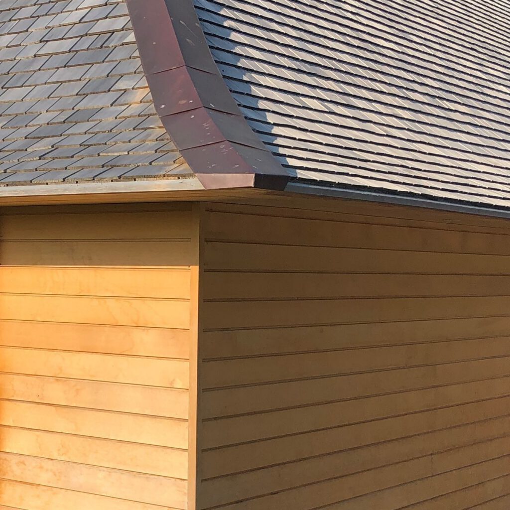 Close-up of the corner of a building with tan horizontal wooden siding and a shingled roof with a metal ridge cap. Sunlight casts shadows, highlighting the textures of the wood and shingles.