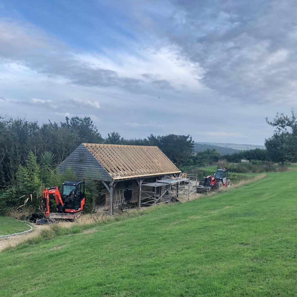 A wooden structure under construction with scaffolding around it, two small excavators nearby, and a grassy field in the foreground under a cloudy sky.