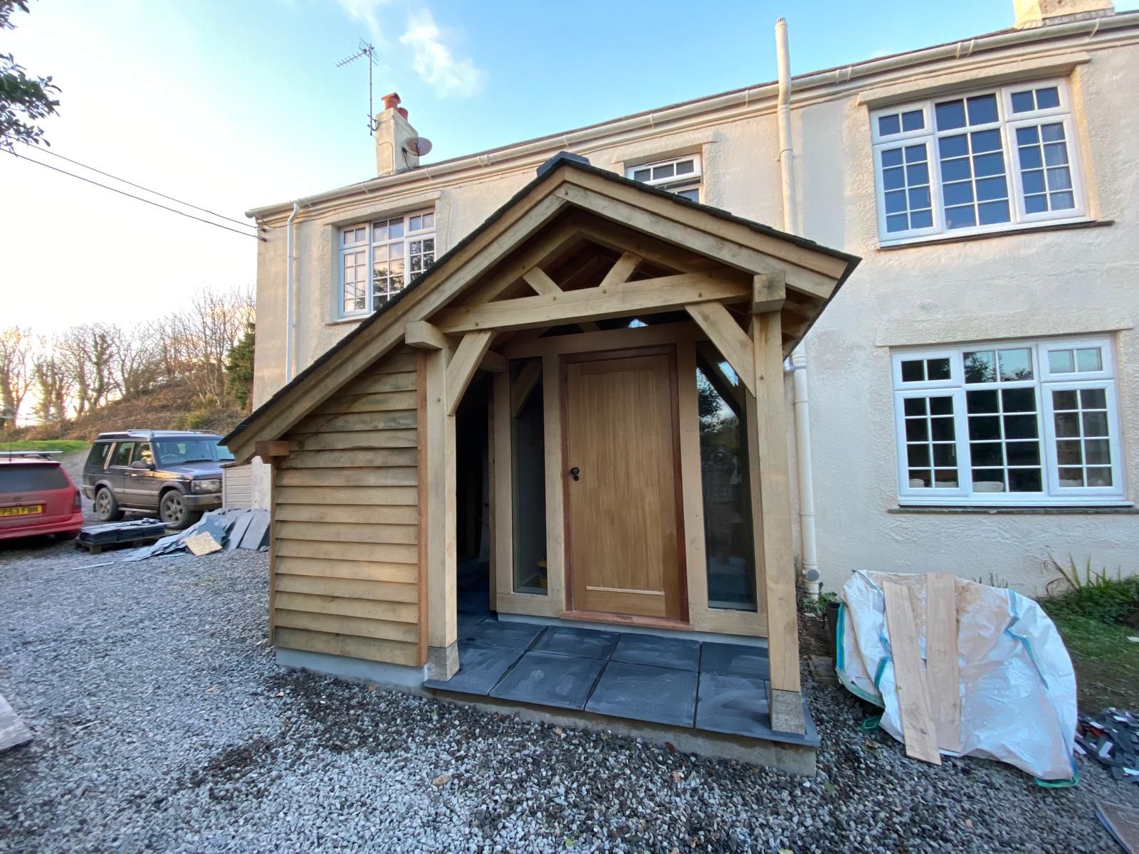 A wooden porch with a pitched roof and tiled floor is attached to the entrance of a cream-colored house; cars and building materials are visible on a gravel driveway nearby.