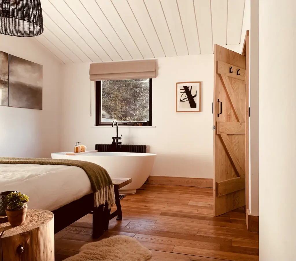 A cozy bedroom features a freestanding bathtub near a window, a wooden floor, a bed with white linens and a green throw, a small wood side table, and a barn-style door partially open. Minimalist decor and warm lighting.