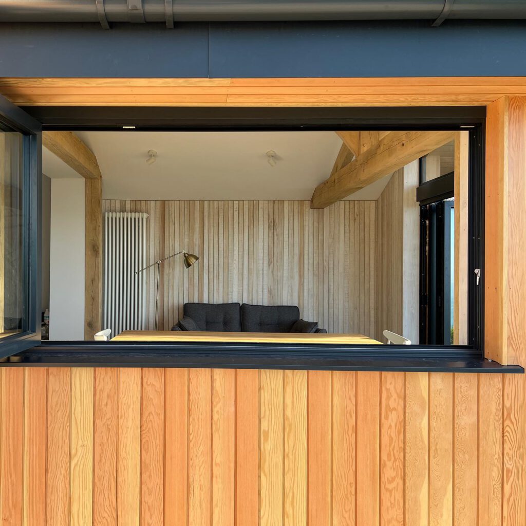 A large open window in a wooden-paneled wall reveals a cozy living room with a gray sofa, a yellow table, a radiator, and a floor lamp against light wood interior walls and exposed beams.