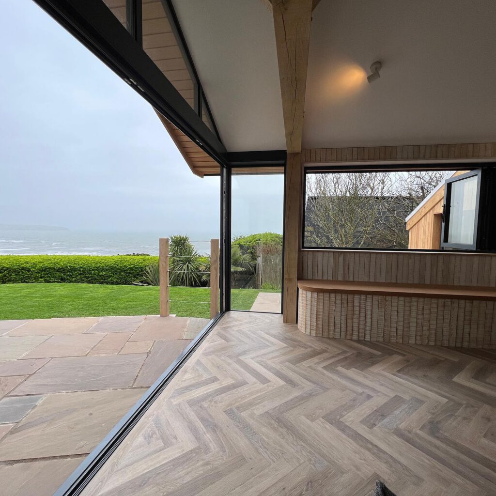Modern room with herringbone wood flooring and large glass doors opening to a stone patio, green lawn, and ocean view on a cloudy day. A window seat and louvered walls are visible inside.
