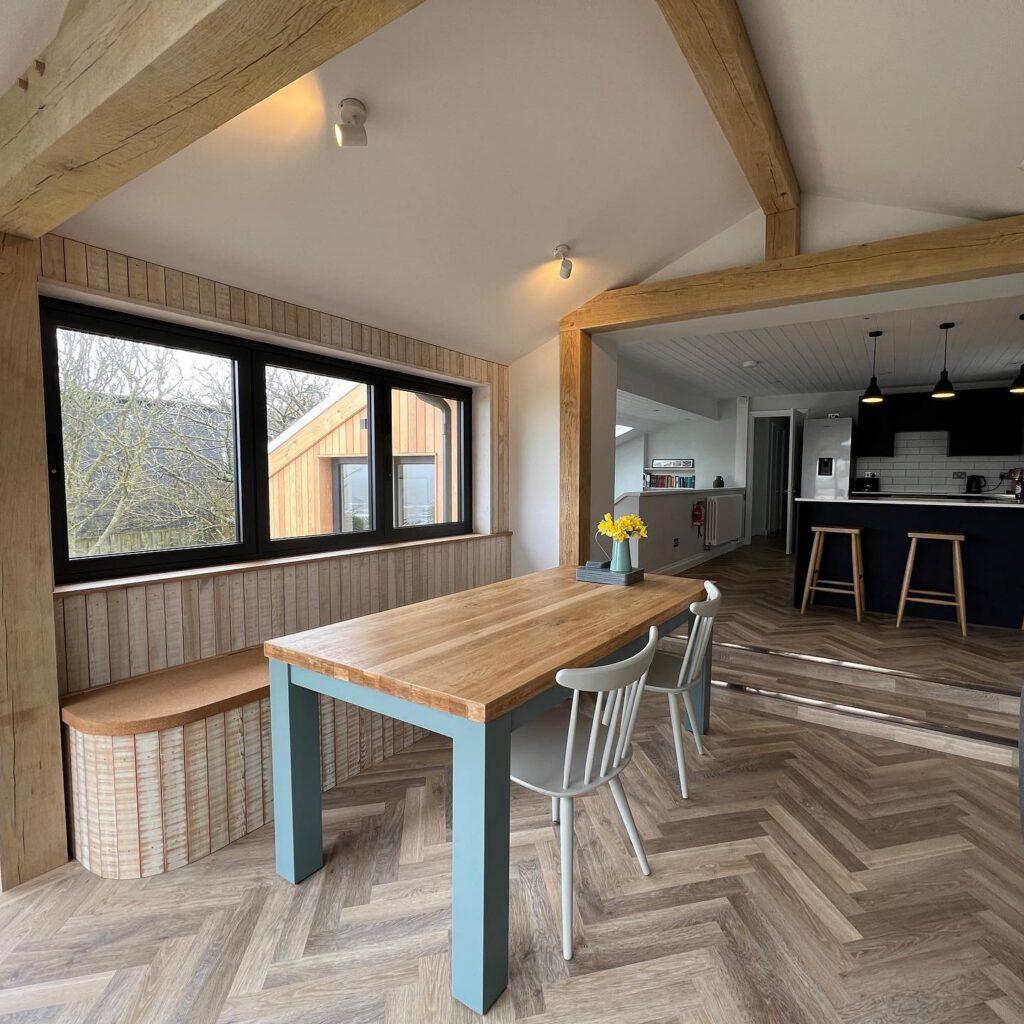 A bright, modern dining area with a wooden table, two chairs, a bench, large windows, and a view into a sleek kitchen. Natural light fills the space, highlighting wood beams and herringbone-patterned flooring.