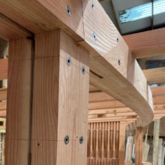 Close-up of a wooden beam joint in a timber frame structure, showing precisely cut wood and metal fasteners securing the connection. Natural light filters in from a nearby window in the workshop.