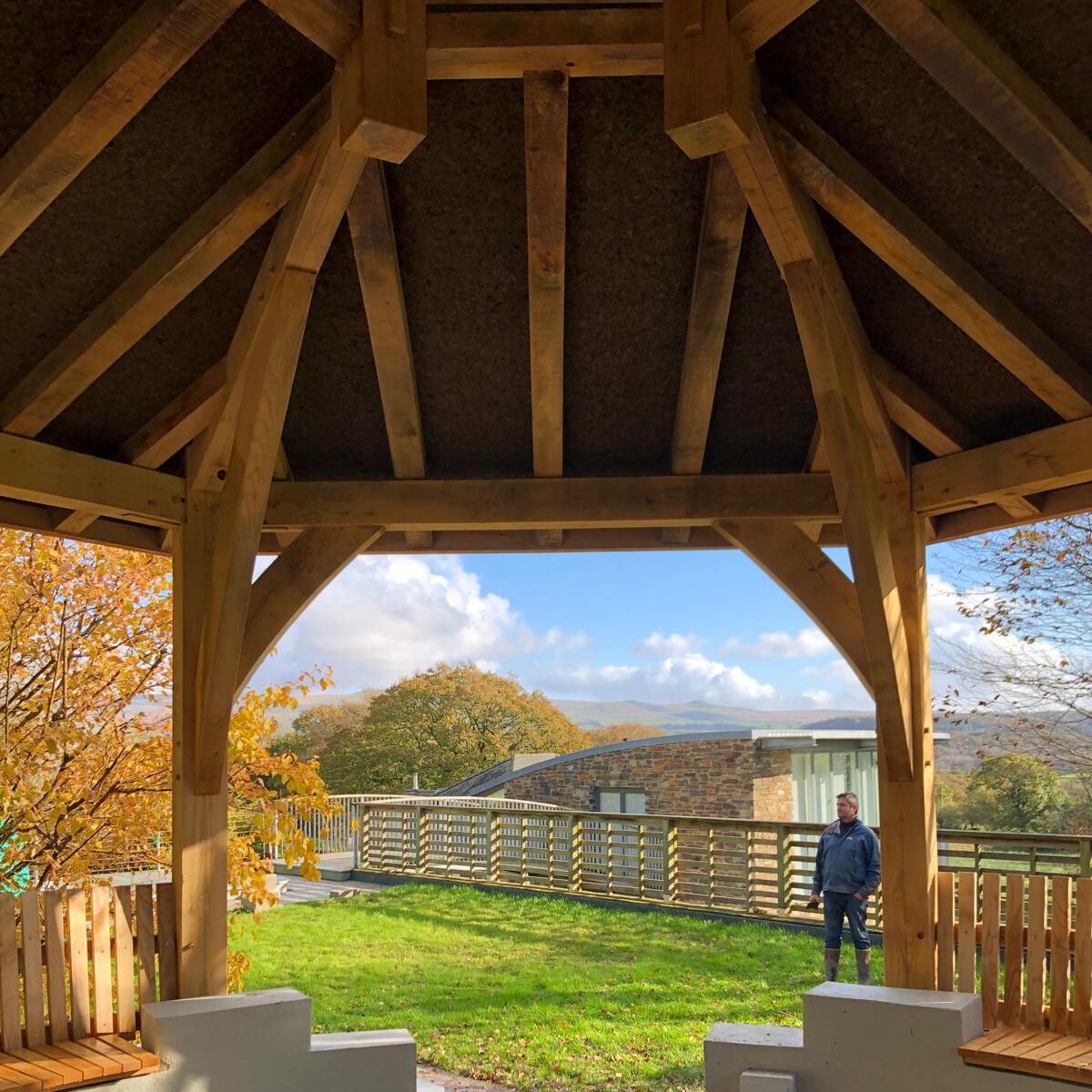 View from inside a wooden gazebo looking out onto a grassy area with autumn trees, a wooden fence, and a person standing outside. The sky is blue with some clouds.