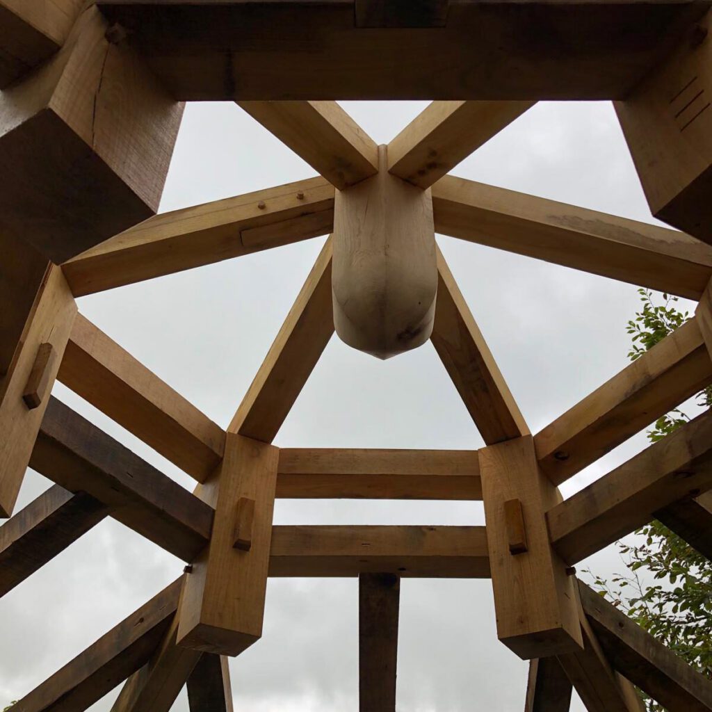 View looking up at the wooden beams and joints of a timber-framed roof structure, with a cloudy sky visible through the open framework.