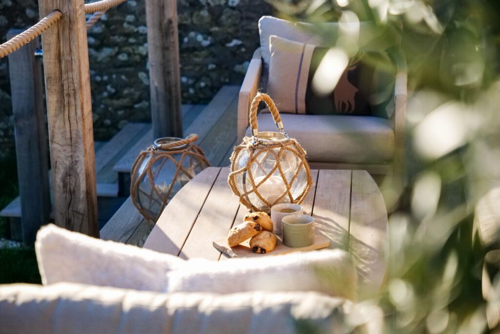 A cozy outdoor seating area with a wooden table holding two rope lanterns, a plate of pastries, and a mug. Cushioned seats and stairs with rope railing are in the background, with sunlight softly illuminating the scene.