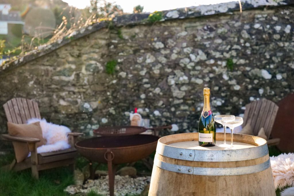A bottle of champagne and two glasses sit on a wooden barrel in a rustic garden with wooden chairs, a fire pit, and a stone wall in the background. Sunlight adds a warm, inviting atmosphere.