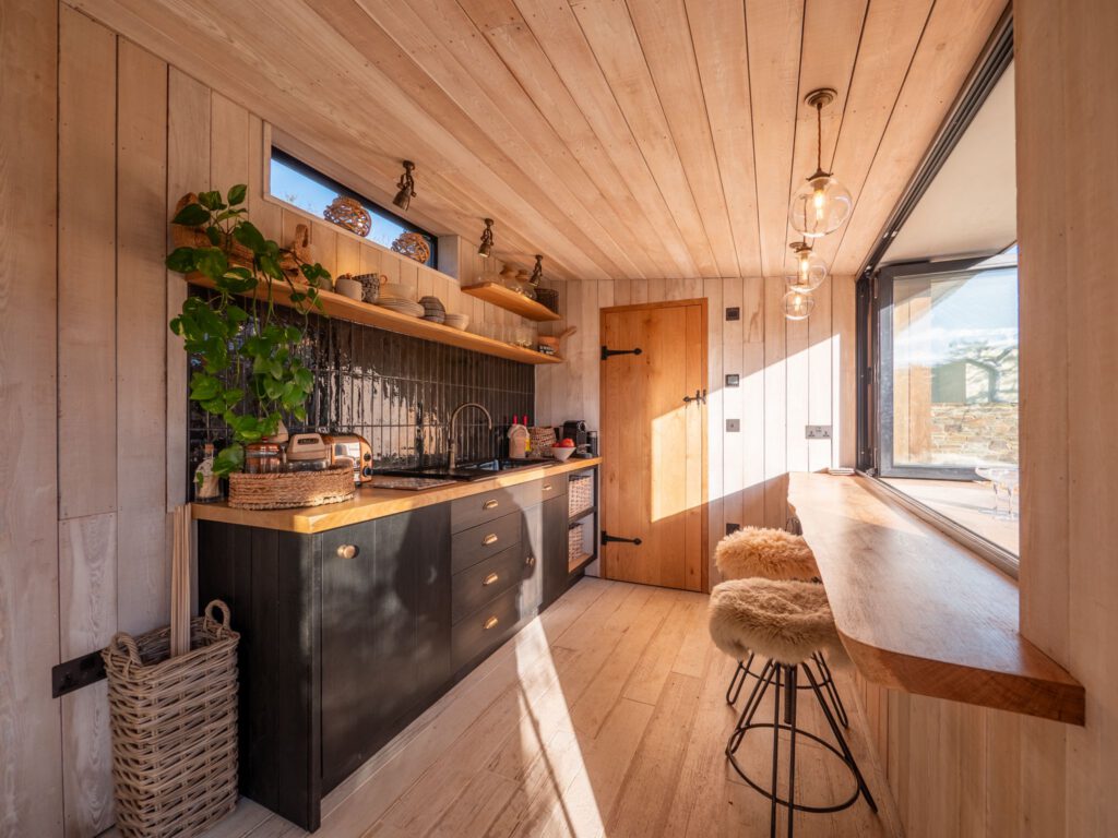 A bright, modern kitchen with wood-paneled walls and ceiling, open shelving, black cabinetry, a large window, hanging lights, potted plant, and two fur-topped bar stools at a wooden counter. Sunlight streams in from the window.