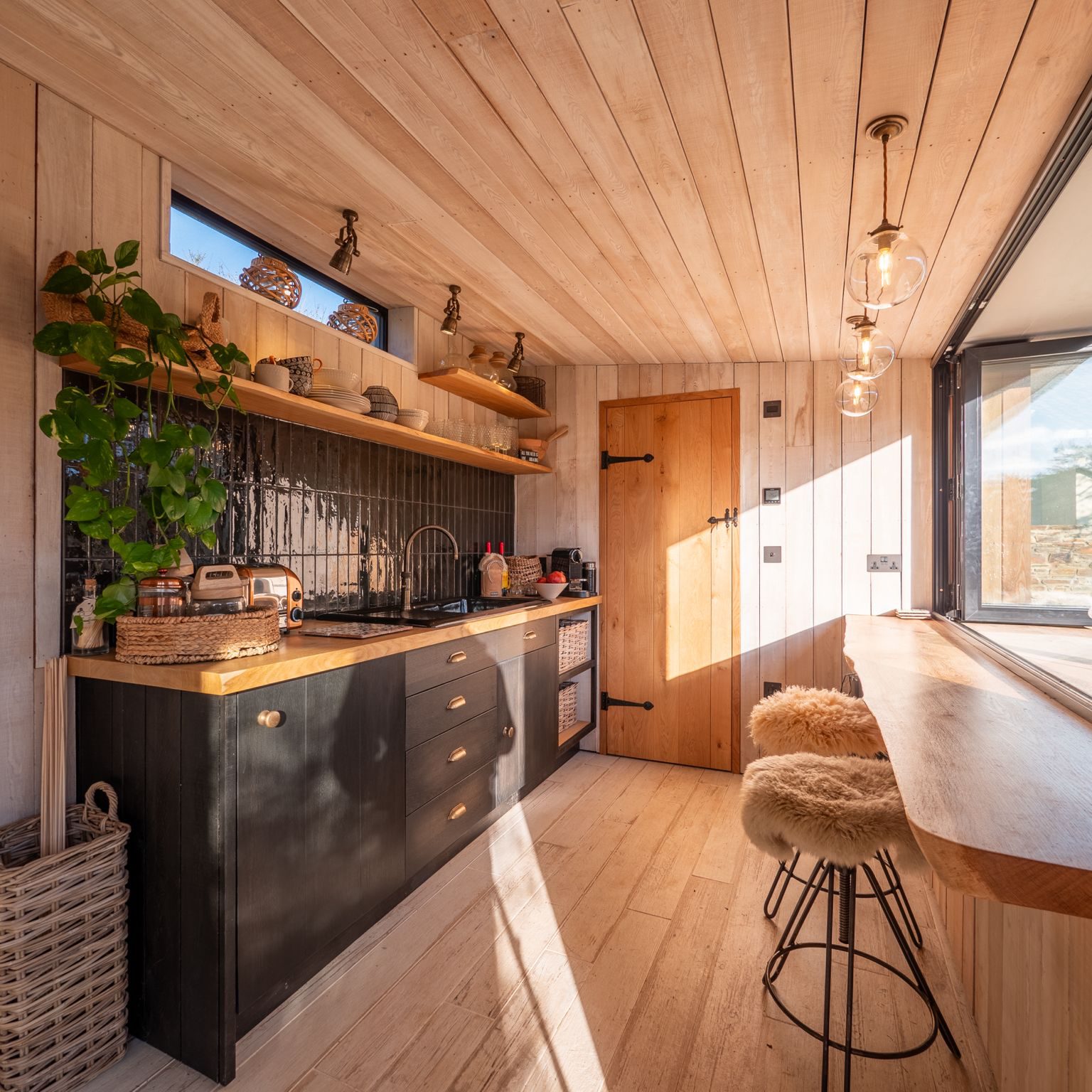 A cozy, sunlit kitchen with wooden floors and ceiling, black cabinets, open shelves, hanging lights, a potted plant, and two bar stools with sheepskin covers at a window-side counter.
