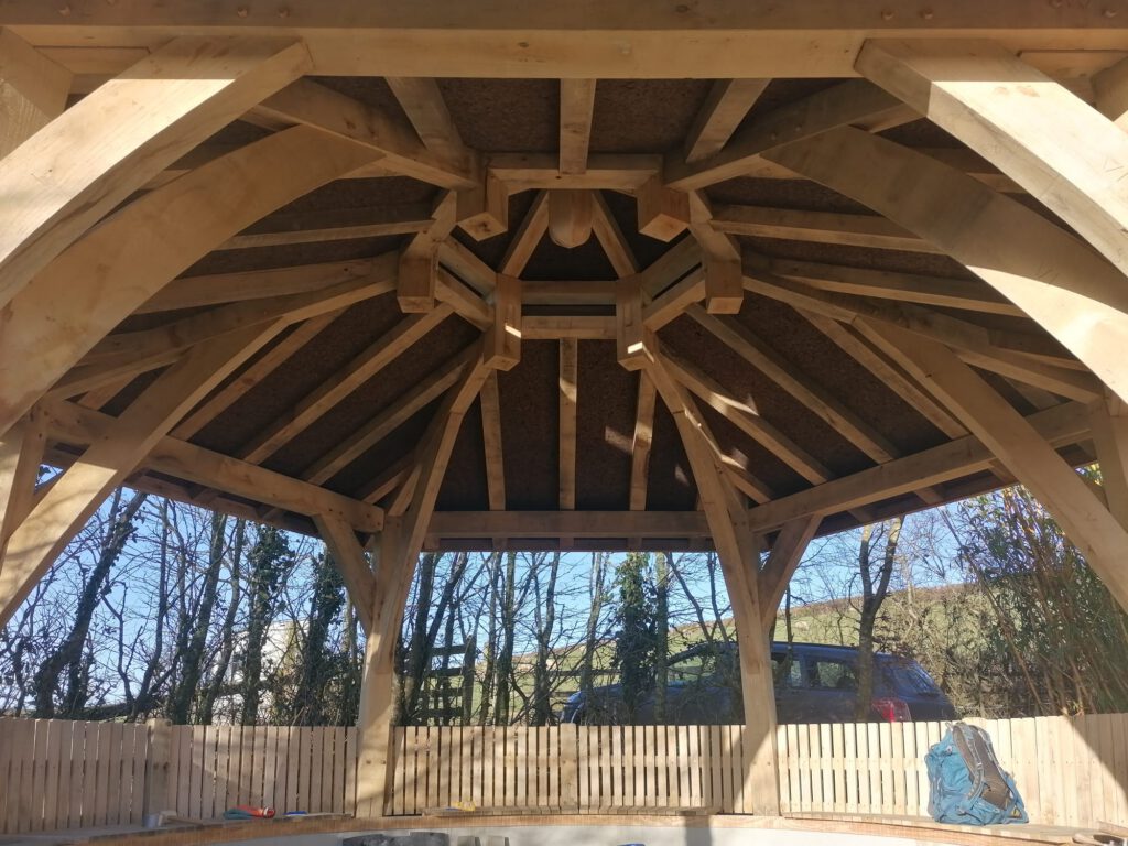 View from inside a wooden gazebo, showing detailed roof beams and open sides. Sunlight filters through the structure, with trees, a wooden fence, a parked car, and a backpack visible outside.