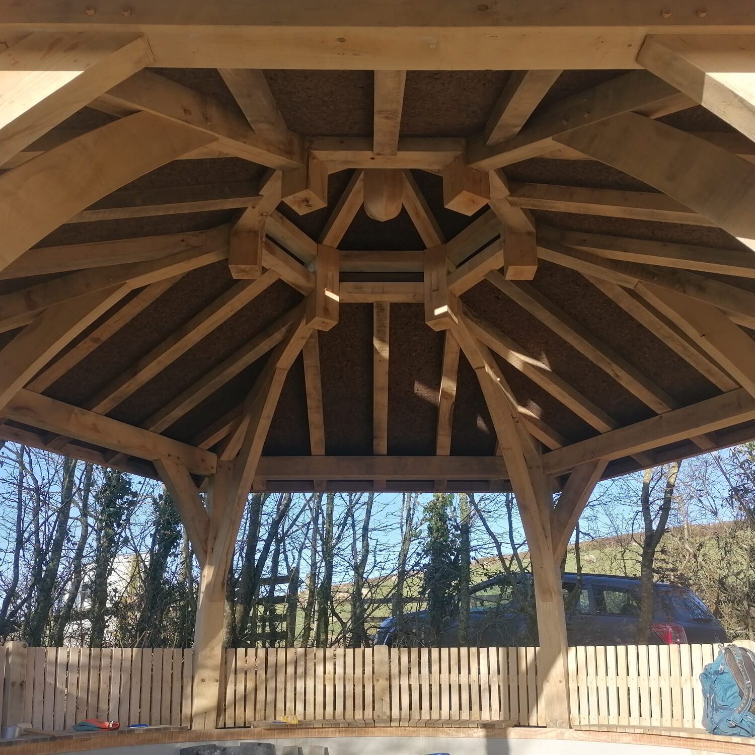 View from inside a wooden gazebo, showing the intricate timber roof structure with beams and supports. Sunlight shines through the open sides, and trees and a wooden fence are visible outside.