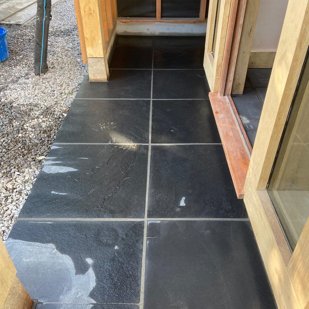 Black tiled patio with some water stains near a wooden-framed glass door, adjacent to a building with wooden beams and gravel on the left side.