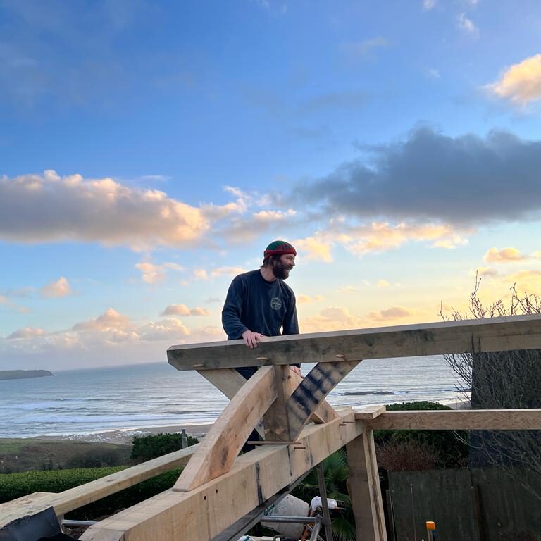 A person wearing a colorful headband stands on a wooden structure under construction, overlooking the ocean and beach with a cloudy sky at sunset in the background.