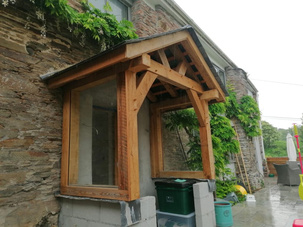 A wooden porch structure with glass sides is attached to a stone house wall. Green plants grow up the wall, and bins and garden tools are placed underneath and nearby. The scene appears to be outdoors on a cloudy day.