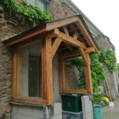 A wooden porch structure with glass sides is attached to a stone house wall. Green plants grow up the wall, and bins and garden tools are placed underneath and nearby. The scene appears to be outdoors on a cloudy day.