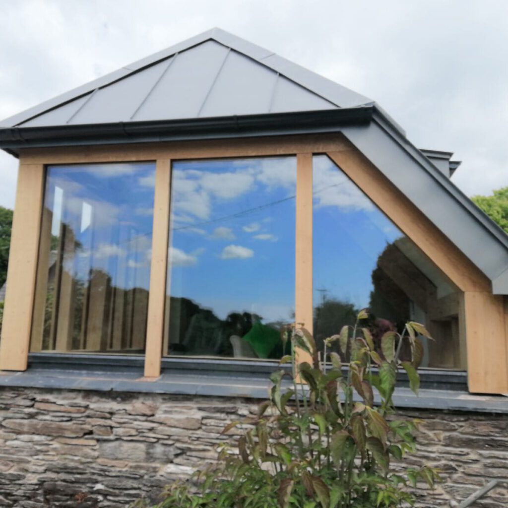 A modern glass extension with a grey metal roof and wooden frame, attached to a stone building, reflecting the blue sky and clouds, with leafy green plants in the foreground.