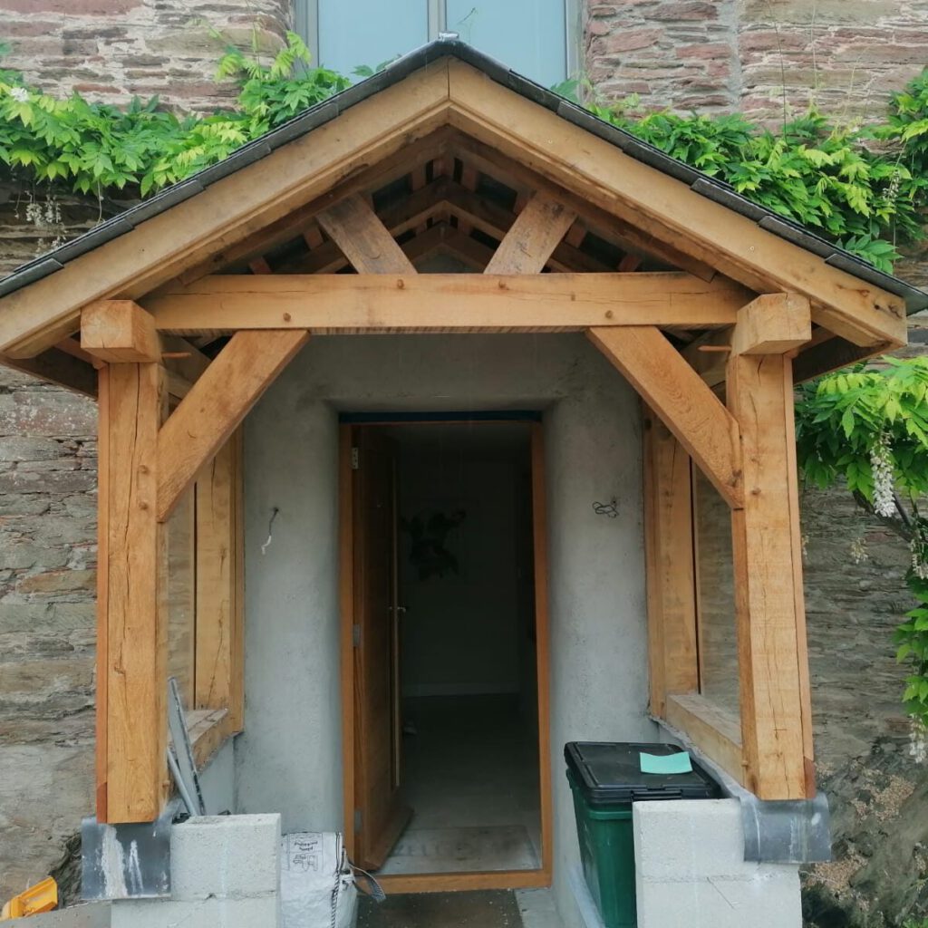A wooden porch with a gabled roof extends from a stone house. The porch has sturdy timber beams and leads to an open doorway. A green bin and cinder blocks sit outside, with climbing plants on the wall above.