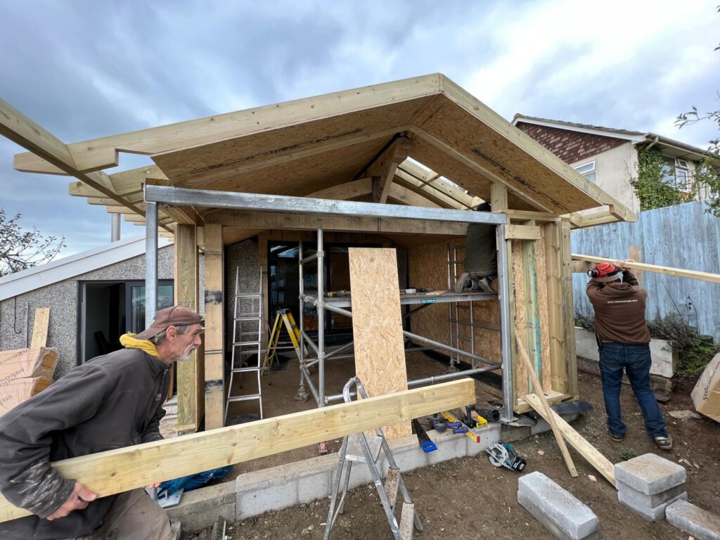 Two men work on the wooden framework of a house under construction, with tools, ladders, and building materials scattered around the site on a cloudy day.