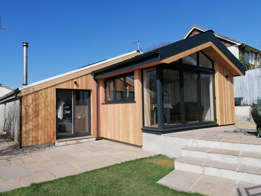 A modern wooden extension with large glass windows and doors, attached to a house. It has a sloped roof and is surrounded by a stone patio and steps, with a green lawn in the foreground under a clear blue sky.