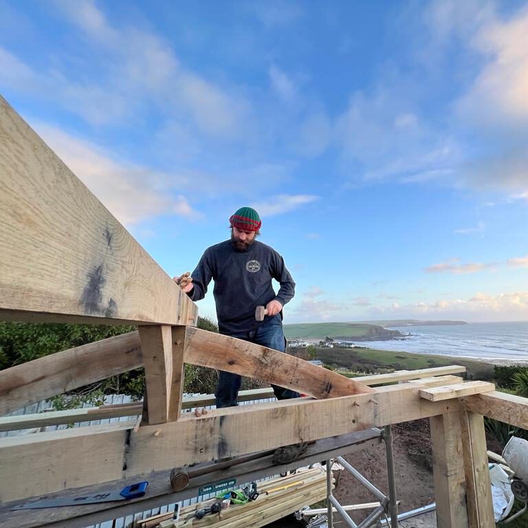 A man wearing a red and green beanie and a dark shirt works on a wooden structure under a blue sky. The scene overlooks a coastline with grassy hills, ocean, and scattered clouds in the background.