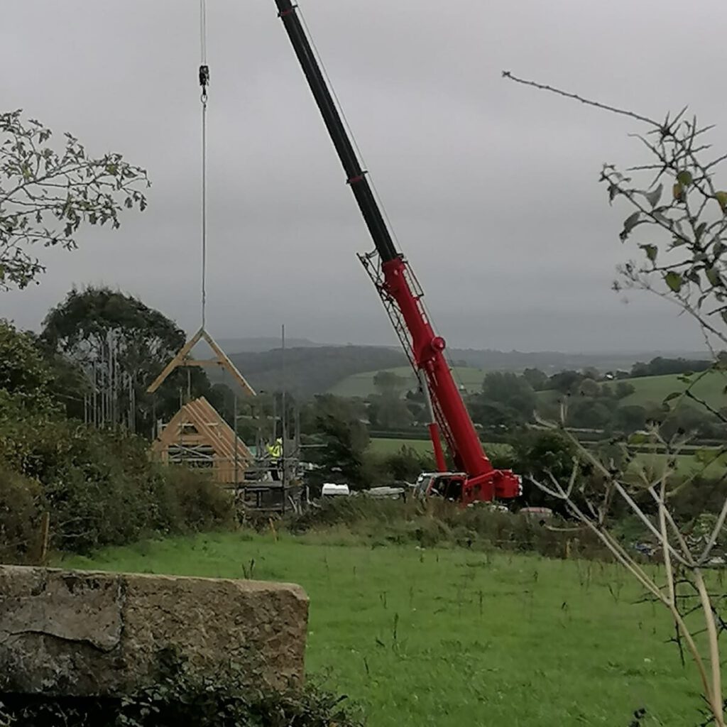 A red crane lifts a wooden house frame at a construction site in a rural area with green fields and rolling hills under an overcast sky.