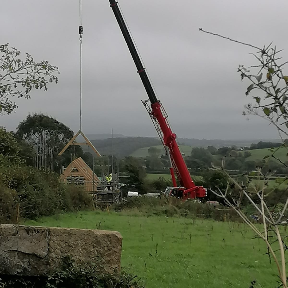 A red crane lifts a wooden house frame at a construction site in a rural area with green fields and rolling hills under an overcast sky.