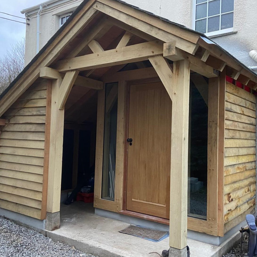 A wooden porch extension with a sloped roof and timber frame stands at the entrance of a house. The porch has vertical wooden panels, a single wooden door, and large windows on either side of the entrance.