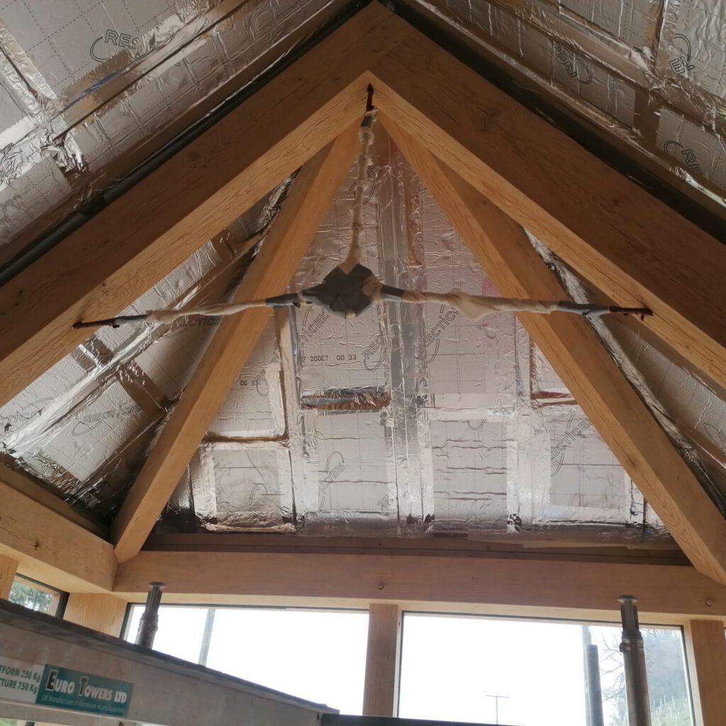 A close-up view of a wooden roof frame with exposed beams and shiny insulation panels; a temporary support made of tape and sticks is fixed between the beams. Scaffolding is visible at the bottom of the image.