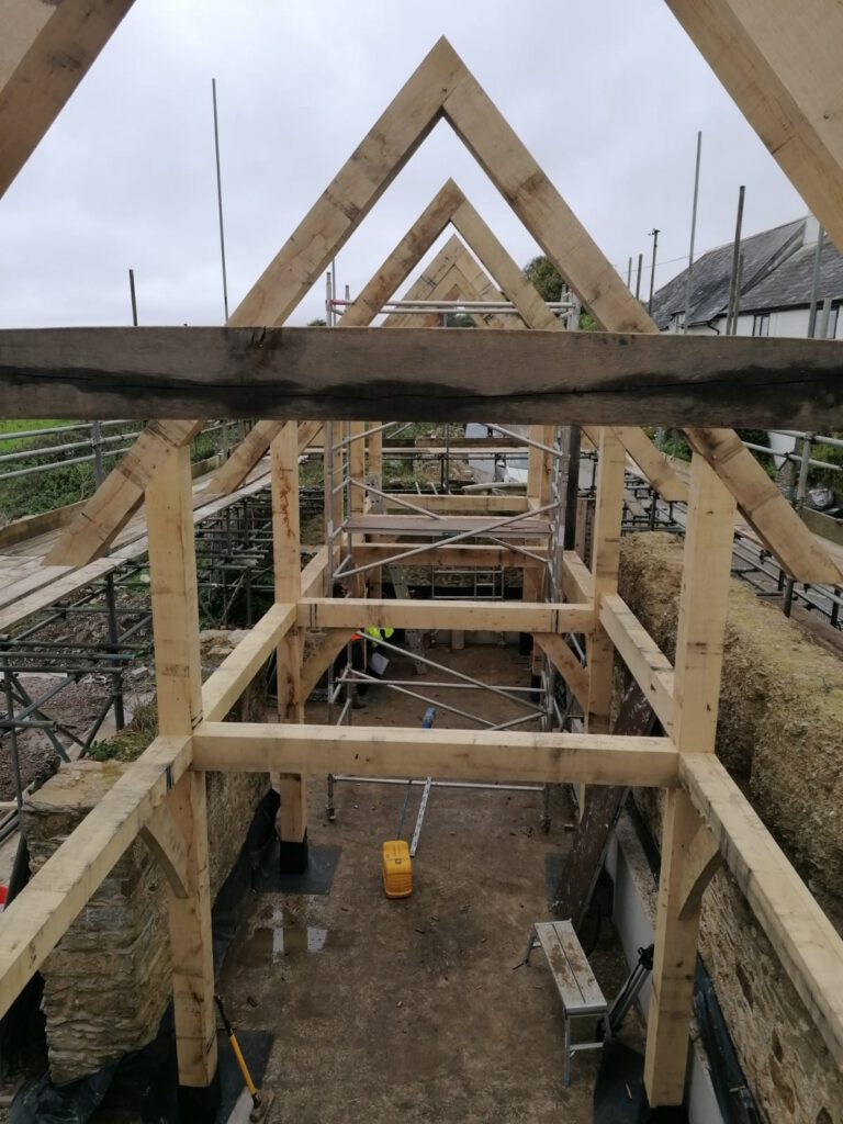 Timber frame structure of a building under construction, surrounded by scaffolding, with exposed beams and tools visible on the ground inside the site.