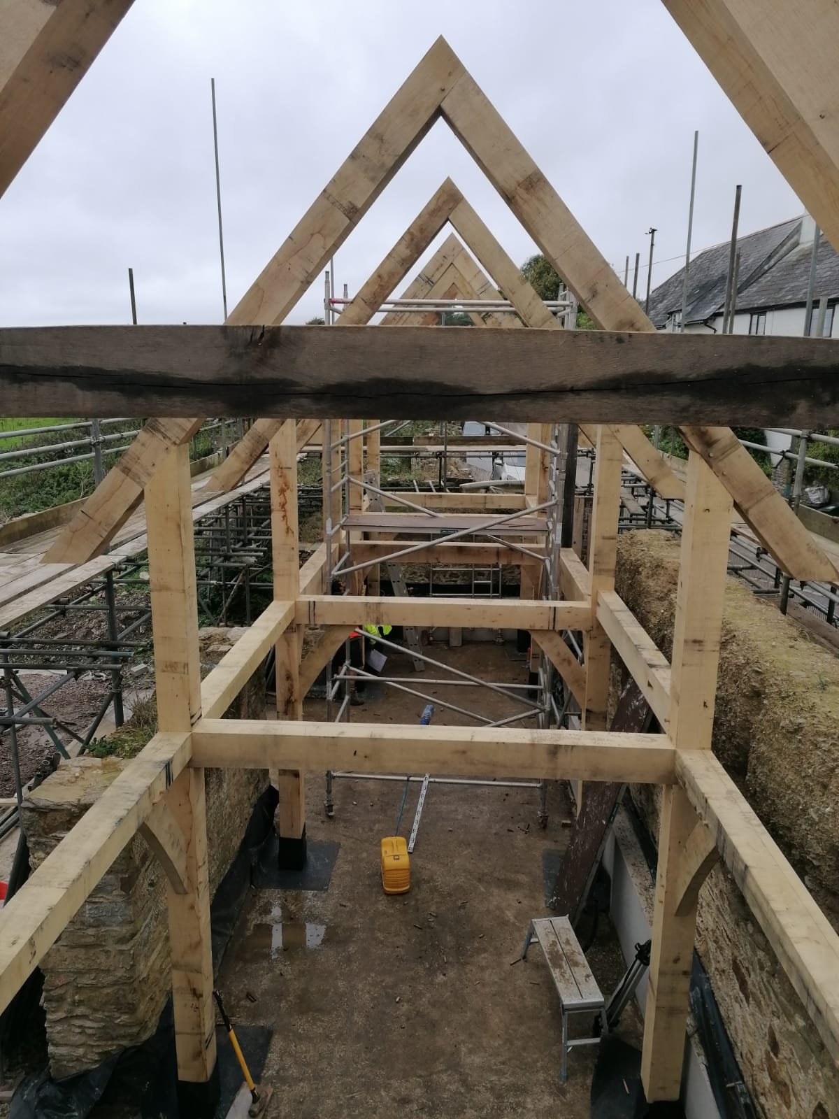 Timber frame structure of a building under construction, surrounded by scaffolding, with exposed beams and tools visible on the ground inside the site.