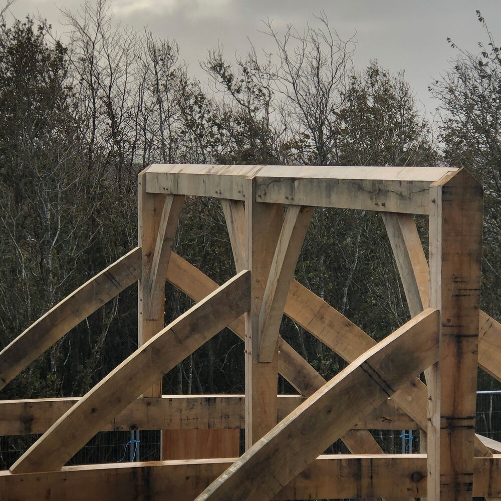 Timber beams and posts form part of a wooden roof frame under construction, with trees and a cloudy sky in the background.