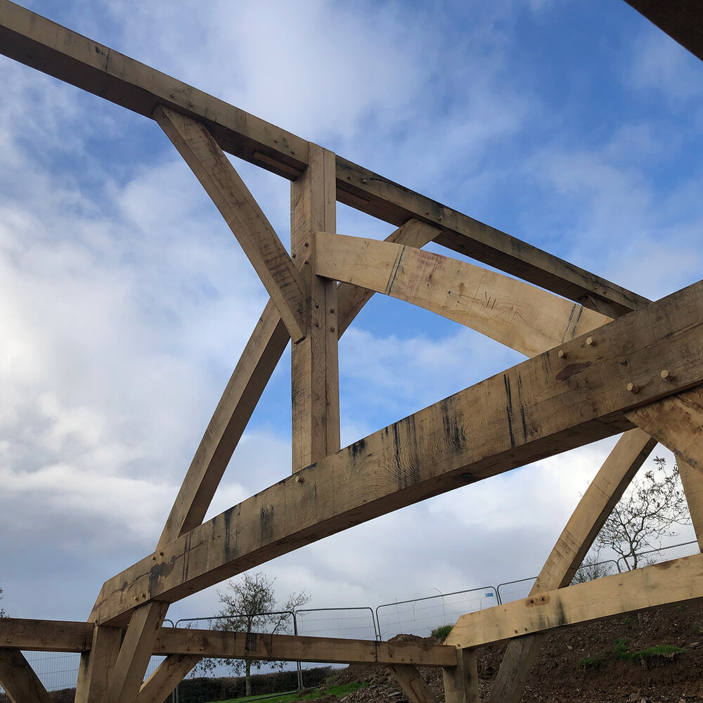 Large wooden beams form an arch and triangular frame structure against a partly cloudy blue sky, with metal fencing and trees visible in the background.