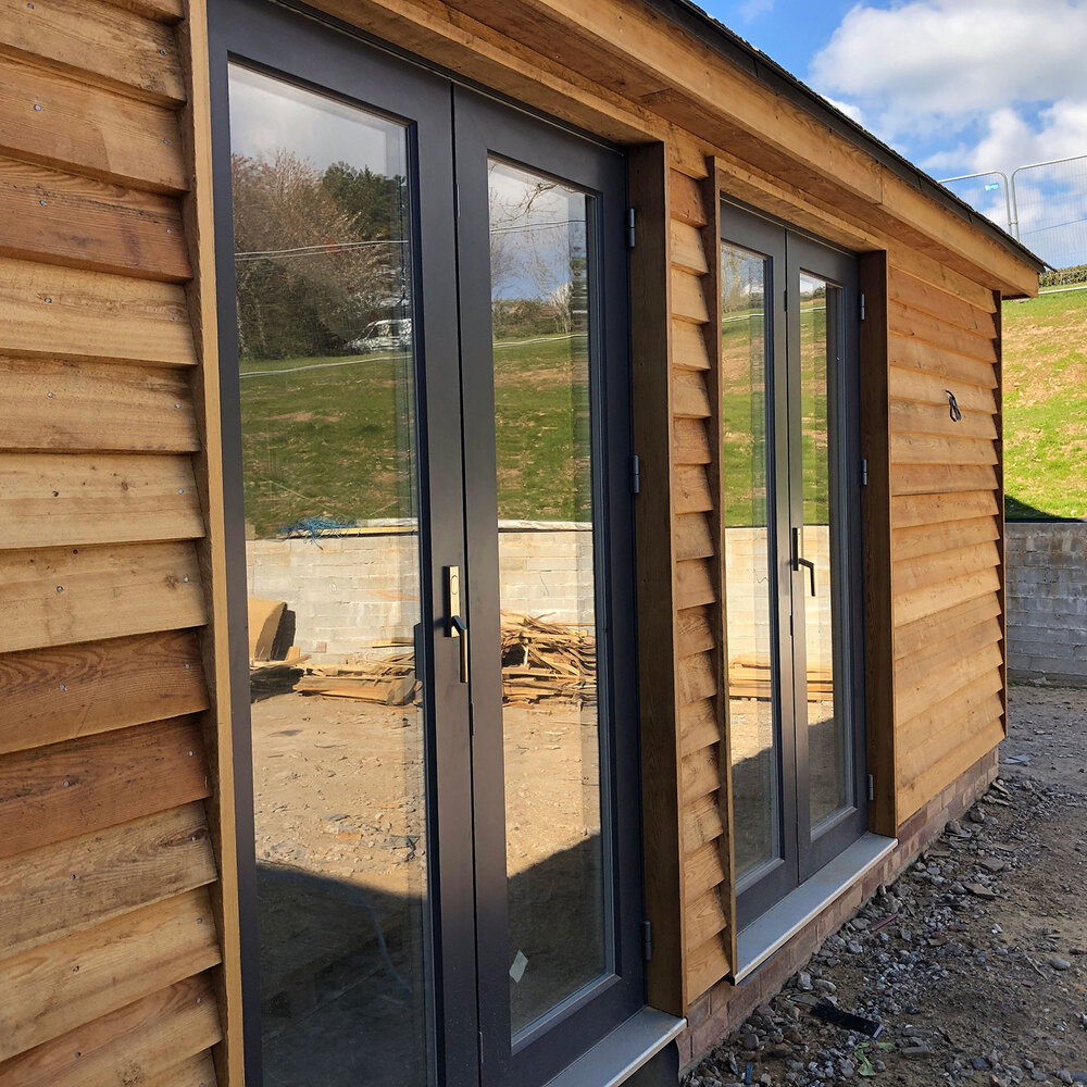 A wooden building with large glass double doors and modern dark frames, reflecting the grassy landscape and blue sky with clouds outside. The ground is rough with some stones and building materials nearby.