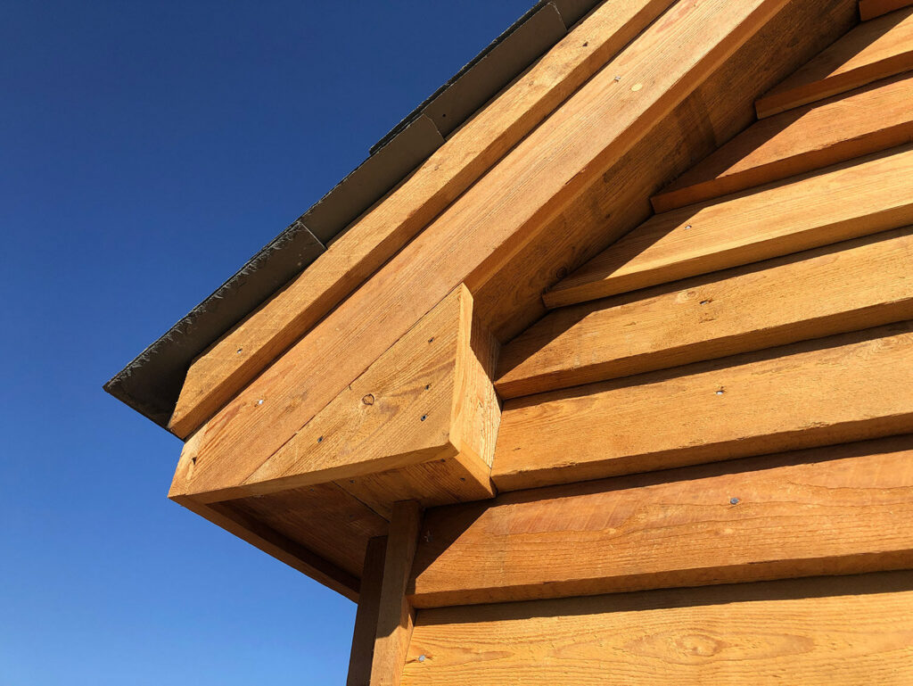 Close-up of a wooden building’s exterior, showing horizontal wooden planks and a gabled roof against a clear blue sky. The wood appears new and is a warm, golden color.