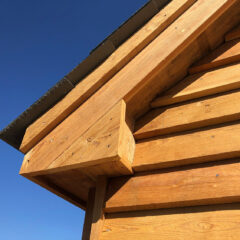 Close-up of a wooden building’s exterior, showing horizontal wooden planks and a gabled roof against a clear blue sky. The wood appears new and is a warm, golden color.